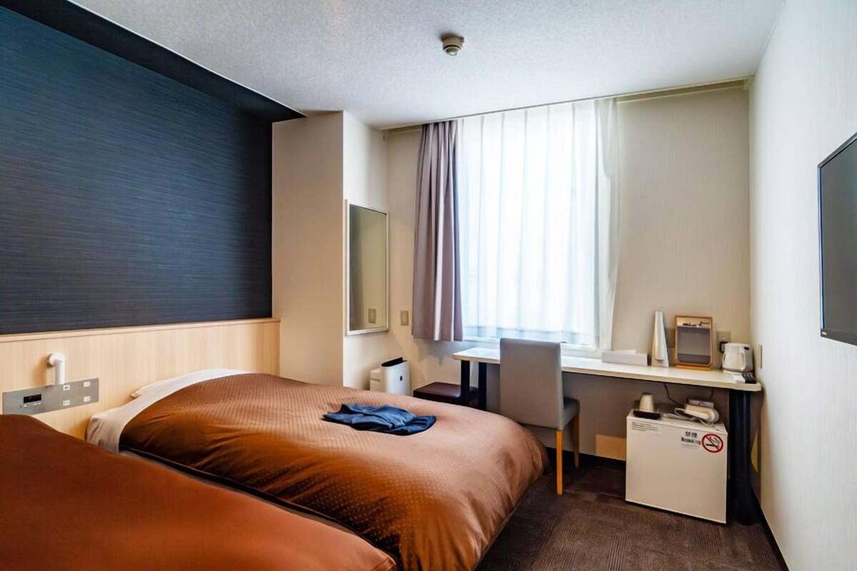 A hotel room with two beds, a desk, a chair, a TV, and a window with curtains.