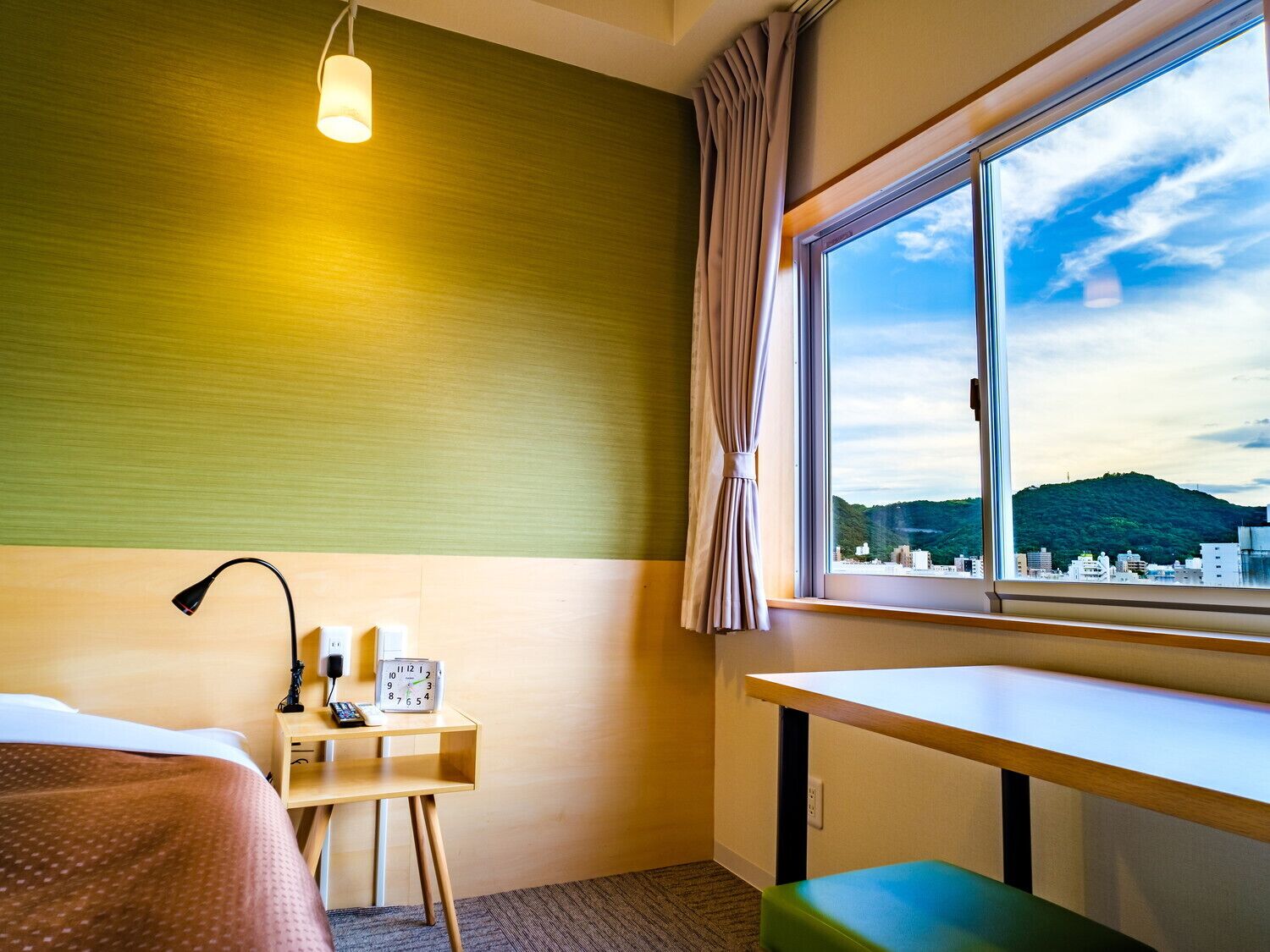 Een hotelkamer met een bed, nachtkastje met een lamp, een klok, een raam met uitzicht op een stadslandschap en heuvels, en een groene bank.