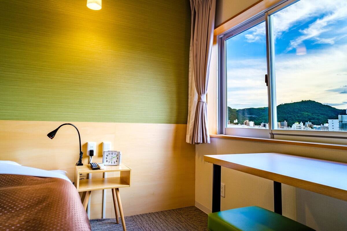 A hotel room with a bed, bedside table with a lamp, a clock, a window with a view of a cityscape and hills, and a green bench.