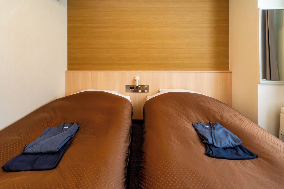 A hotel room with two single beds, a wooden headboard, a nightstand with a lamp, and a window with curtains.