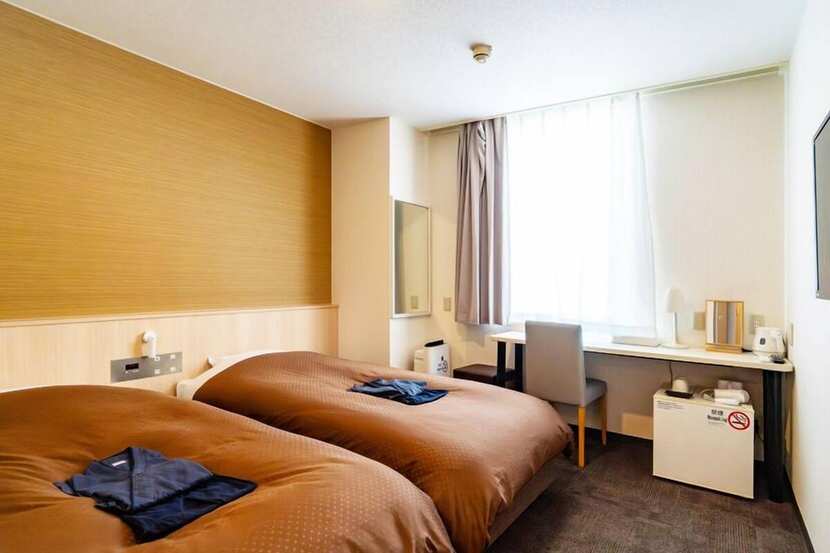 A hotel room with two beds, a desk, a chair, a window with curtains, and a small table with a coffee maker.