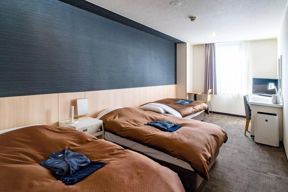 A hotel room with two beds, a desk, a chair, a lamp, and a window with curtains.