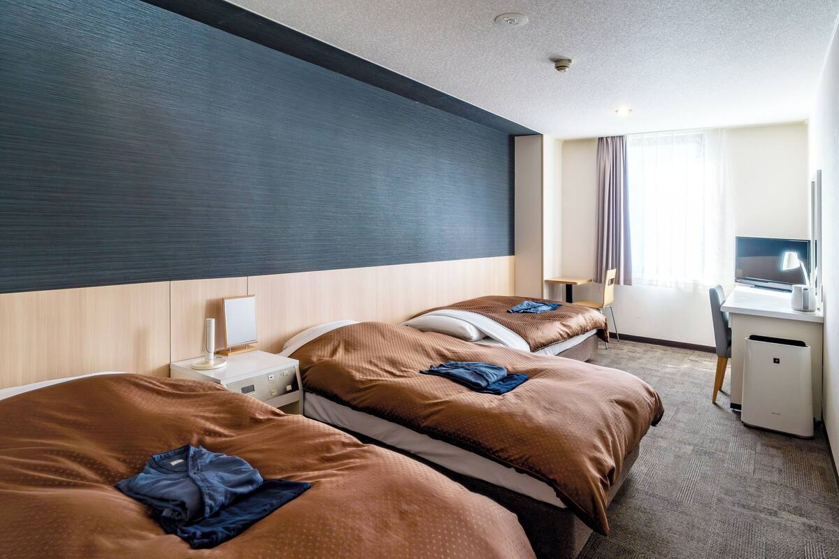 A hotel room with two beds, a desk, a chair, a TV, and a window.
