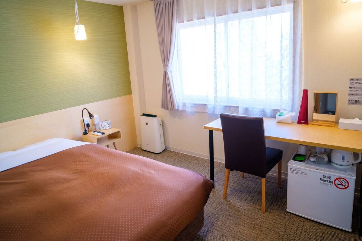 A hotel room with a bed, desk, chair, and a small air purifier.