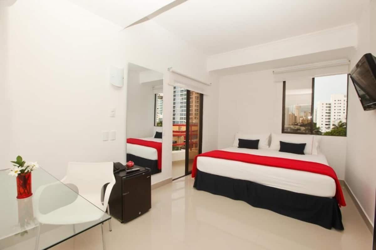 Ein Hotelzimmer mit einem großen Bett, einem Glastisch, einem Sessel und einem an der Wand montierten Fernseher.