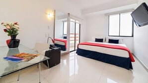 A hotel room with a glass table, a chair, a bed with red and white bedding, a TV mounted on the wall, and a window.