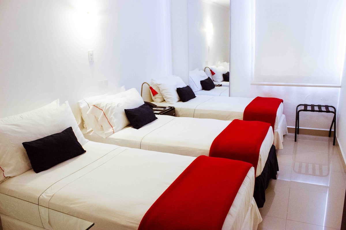 Ein Hotelzimmer mit drei Betten, jeweils mit weißer Bettwäsche und roten Akzenten.