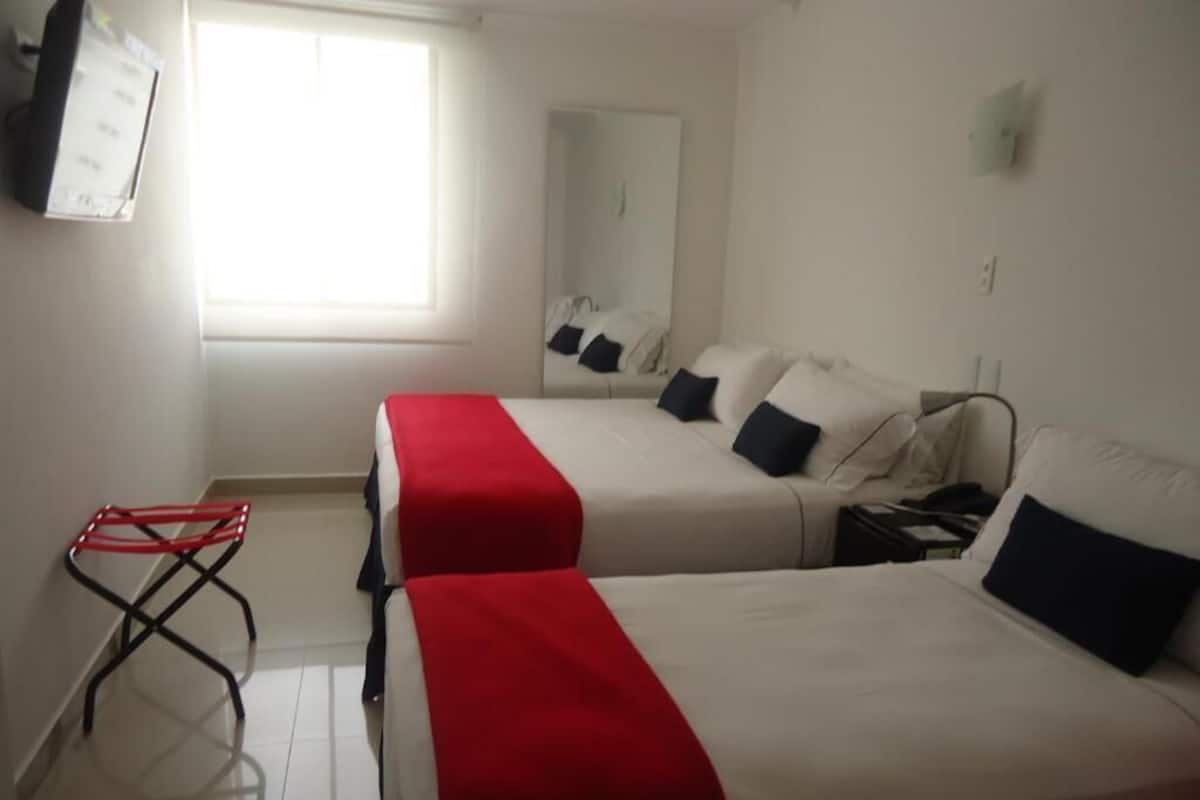 Ein Hotelzimmer mit zwei Betten, einer roten und weißen Bettdecke, einem kleinen Tisch, einem Spiegel und einem Fenster mit Tageslicht.