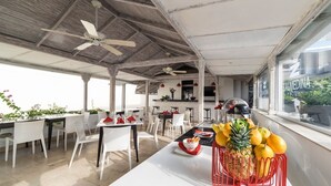A spacious outdoor dining area with white tables and chairs, a ceiling fan, and a view of the hotel building.