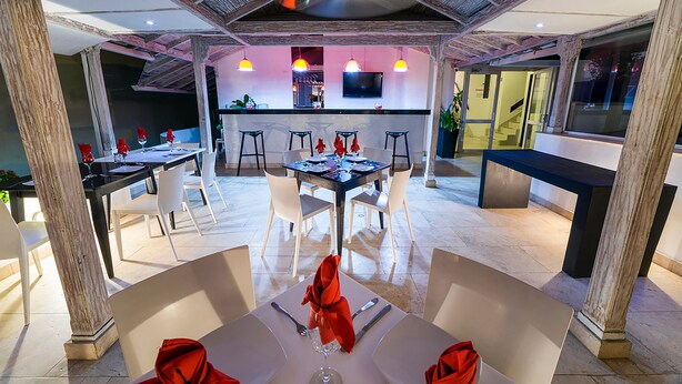 A modern dining area with a high ceiling and exposed beams, featuring a bar and seating with a view of the kitchen and a television.