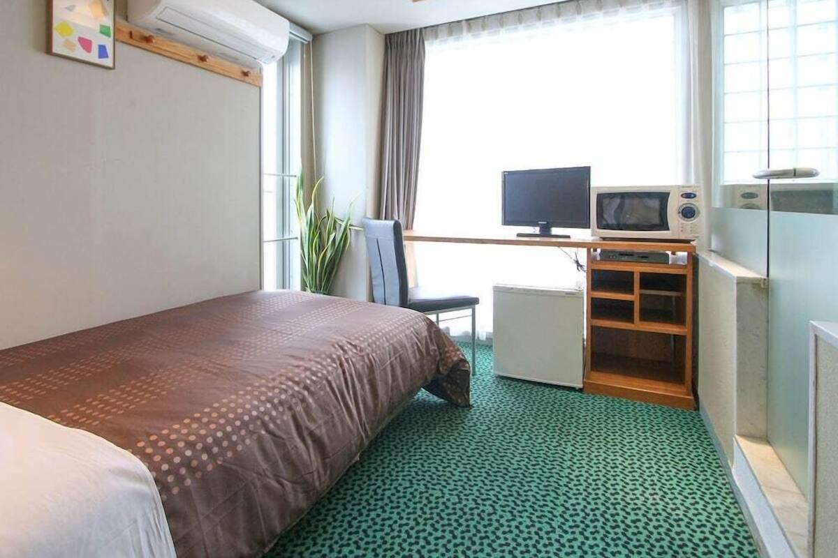 Ett hotellrum med en säng, ett skrivbord med en dator, ett litet kylskåp och ett fönster med gardiner.
