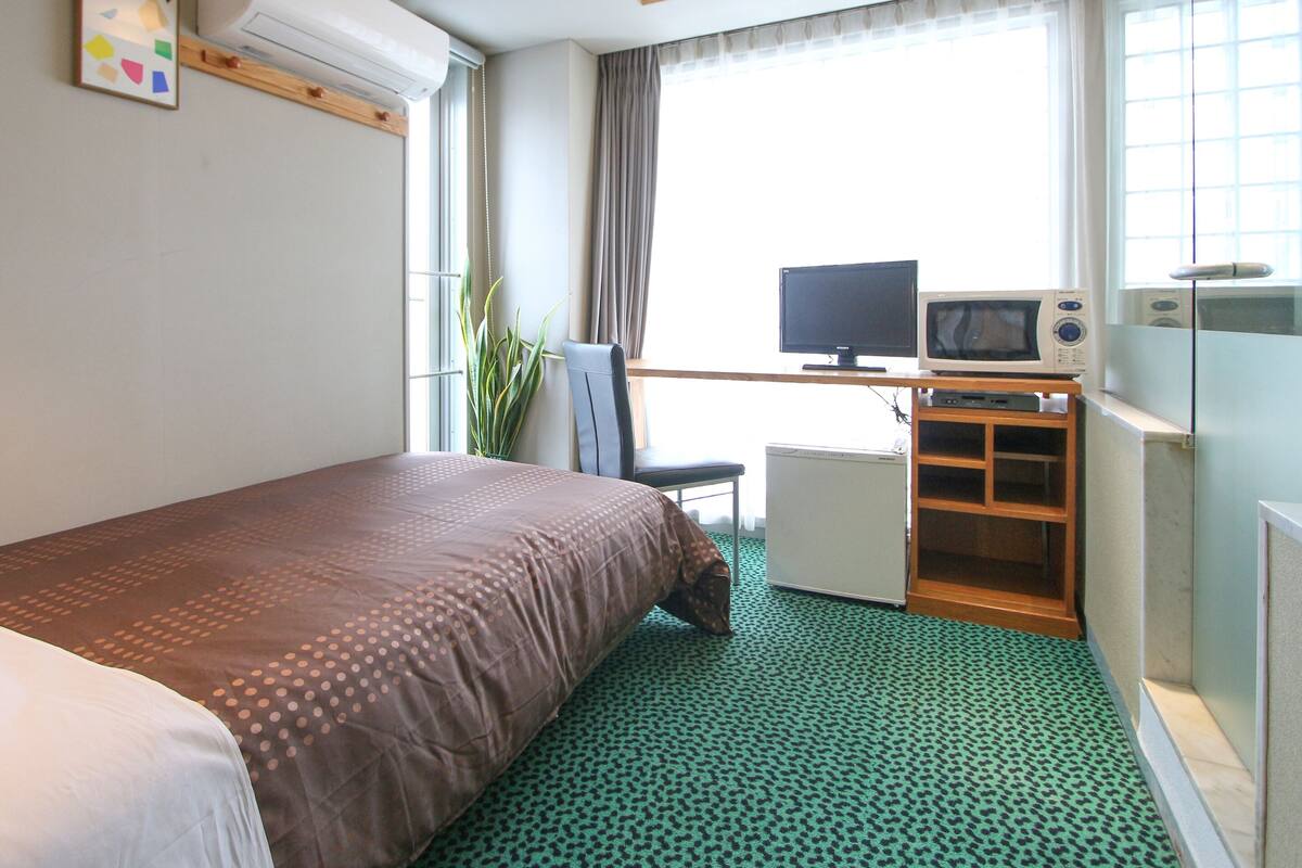 Ett hotellrum med en säng, ett skrivbord med en dator och en mikrovågsugn, ett litet kylskåp och ett fönster med gardiner.