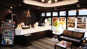 A hotel reception area with a white desk, a black leather sofa, a small table with a candle, and a 'Live Max' magazine rack. - Hotel Livemax Budget Fuchu (Fuchu)