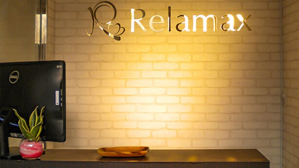 A reception desk with a Dell computer monitor, a potted plant, and a wooden tray. The wall behind features a sign with the word "Relax" illuminated. - Hotel Livemax Budget Fuchu (Fuchu)