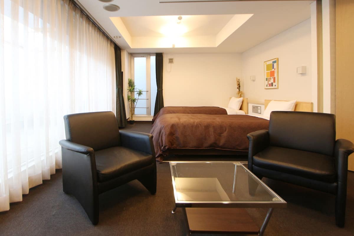 A hotel room with a bed, two armchairs, a glass coffee table, and a large window with curtains.
