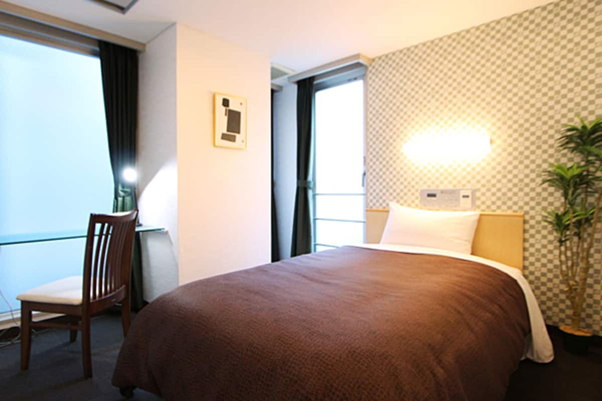 A hotel room with a large bed, a desk with a chair, a window, and a wall-mounted light fixture.