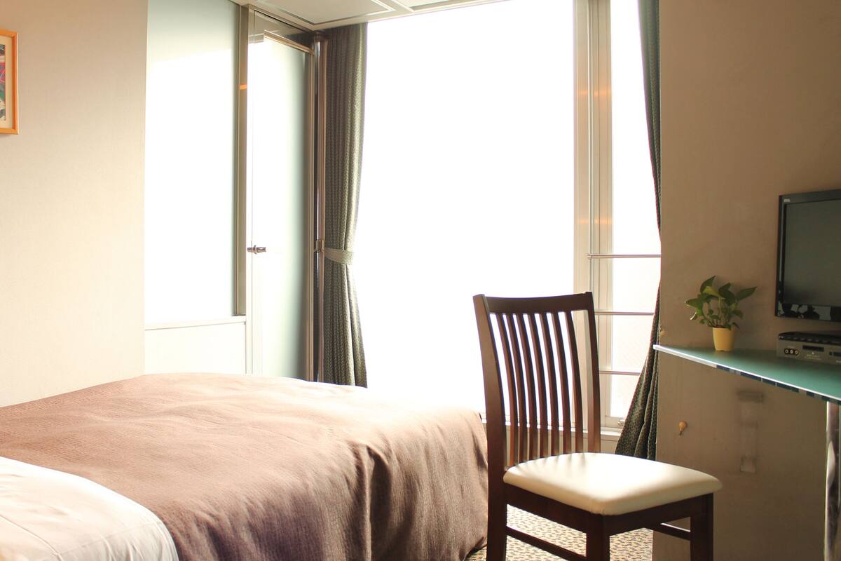 A hotel room with a bed, a desk with a chair, a television, and a window with curtains.