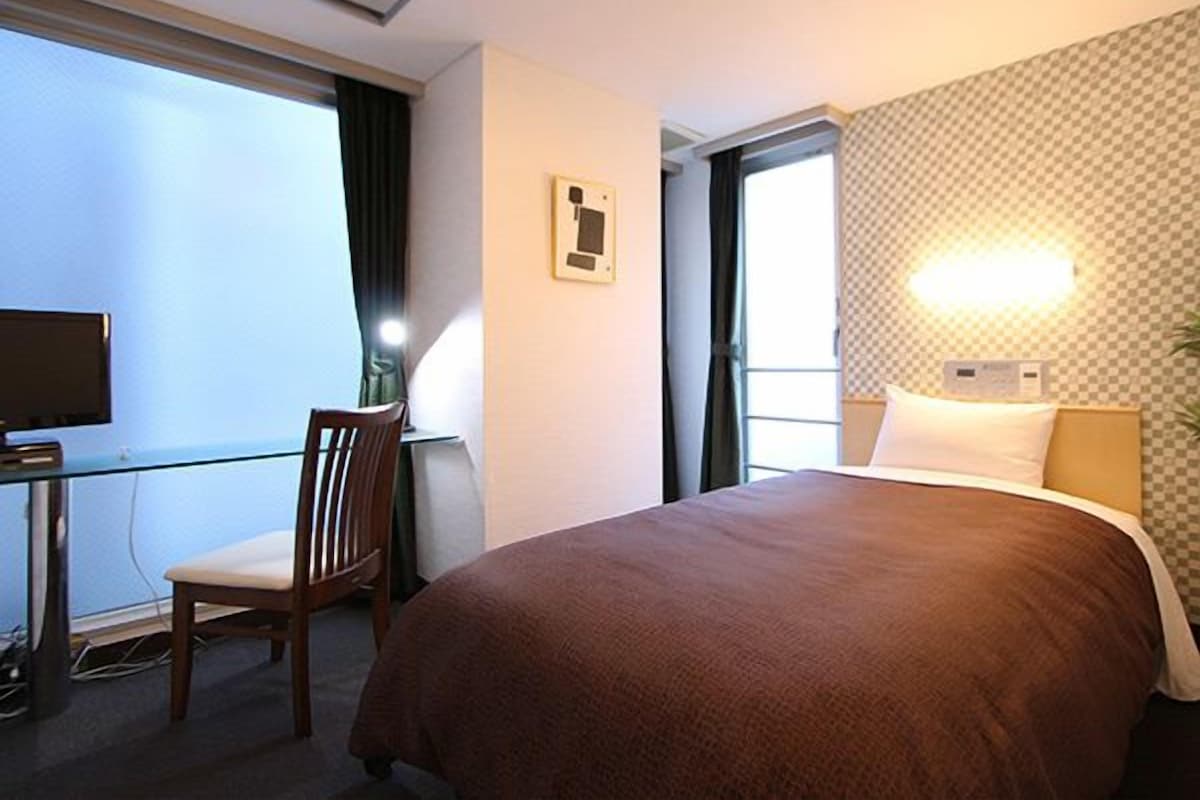 A hotel room with a bed, a desk with a chair, a television, and a window with curtains.