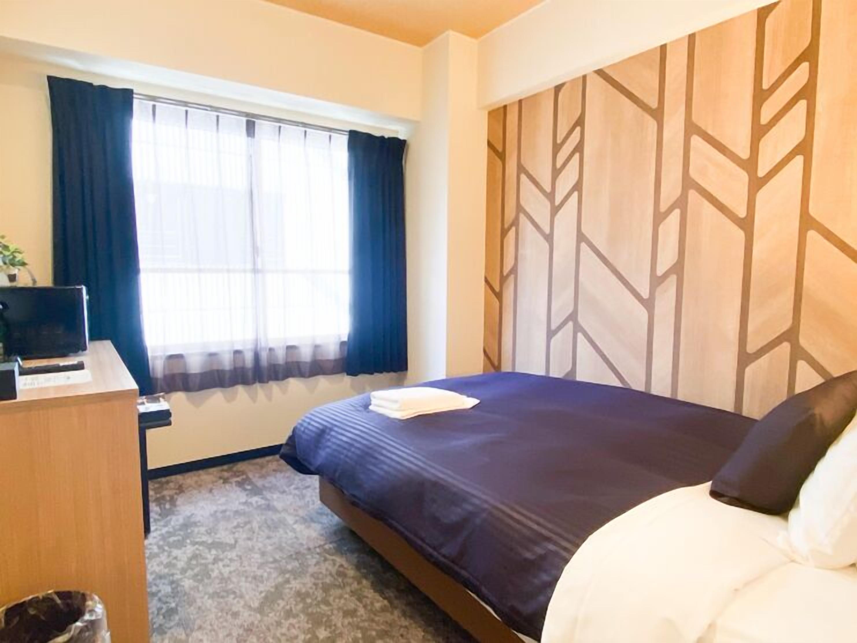 Habitació d'hotel amb un llit, escriptori, cadira, finestra amb cortines i un mur amb un disseny decoratiu.