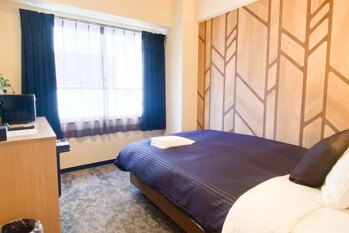 A hotel room with a bed, a desk, a chair, a window with curtains, and a patterned wall.