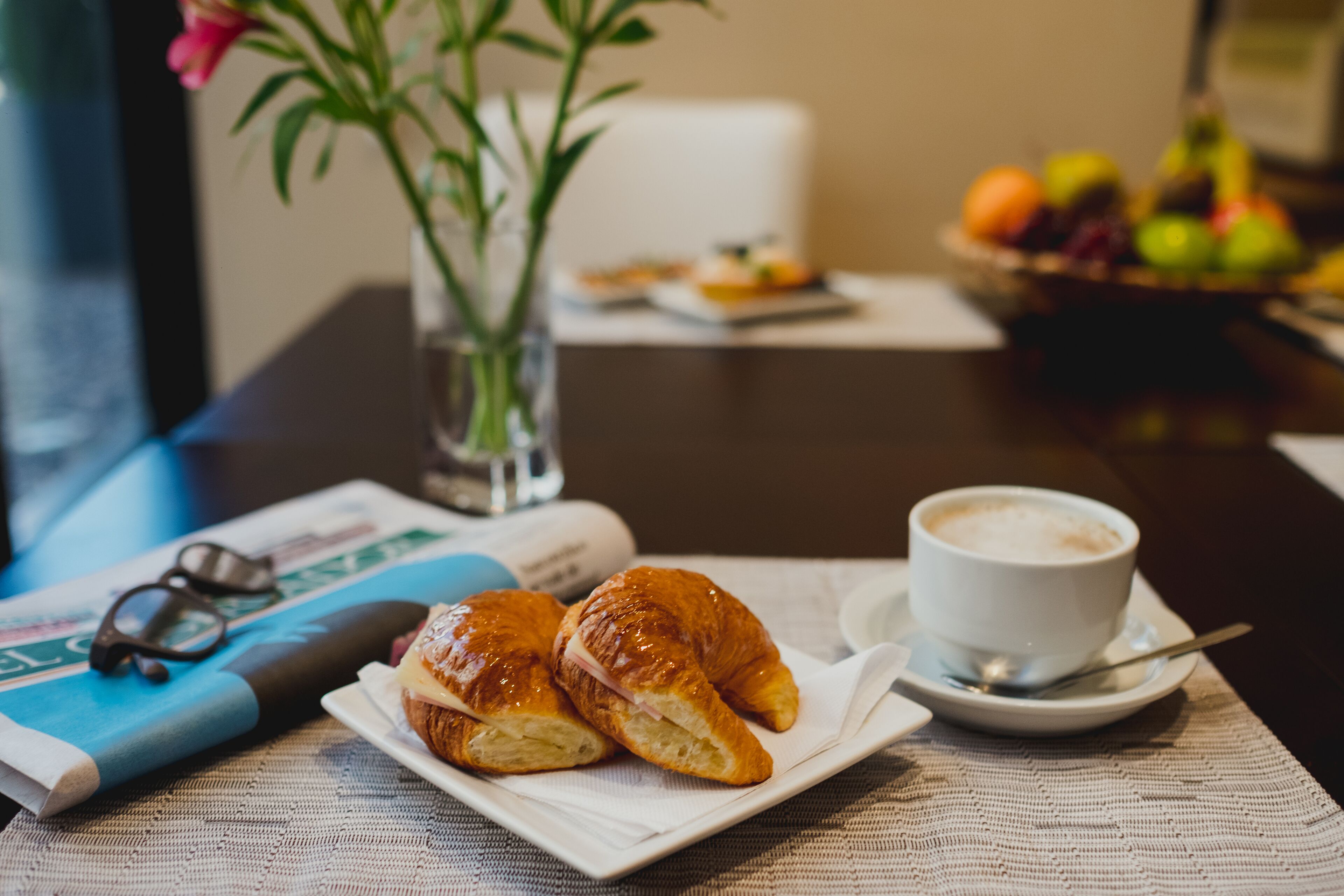 Uma mesa com croissants, uma chávena de café e uma taça de fruta para o pequeno-almoço.