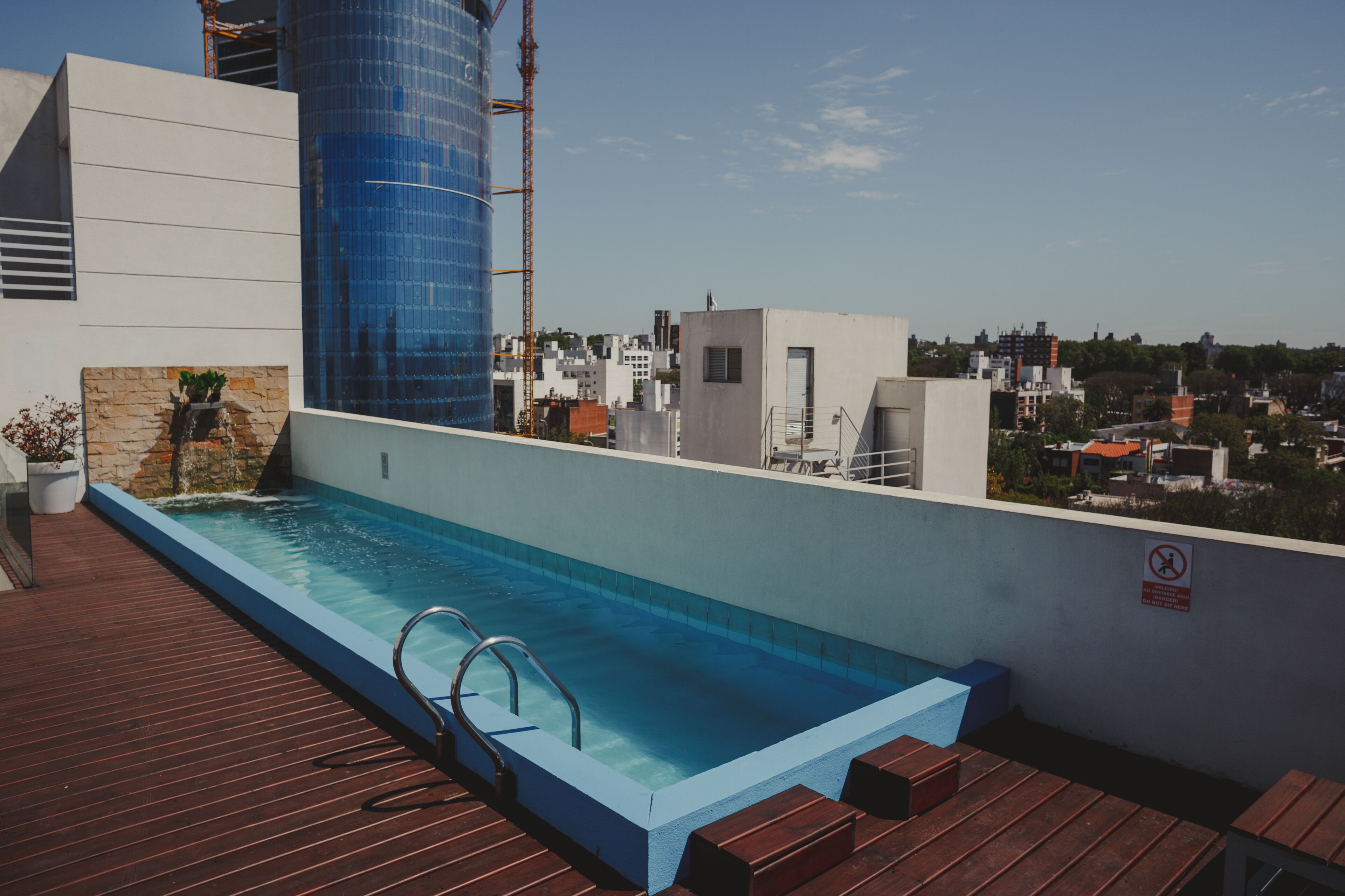 A rooftop swimming pool with a wooden deck, a blue-tiled pool, and a cityscape view in the background.