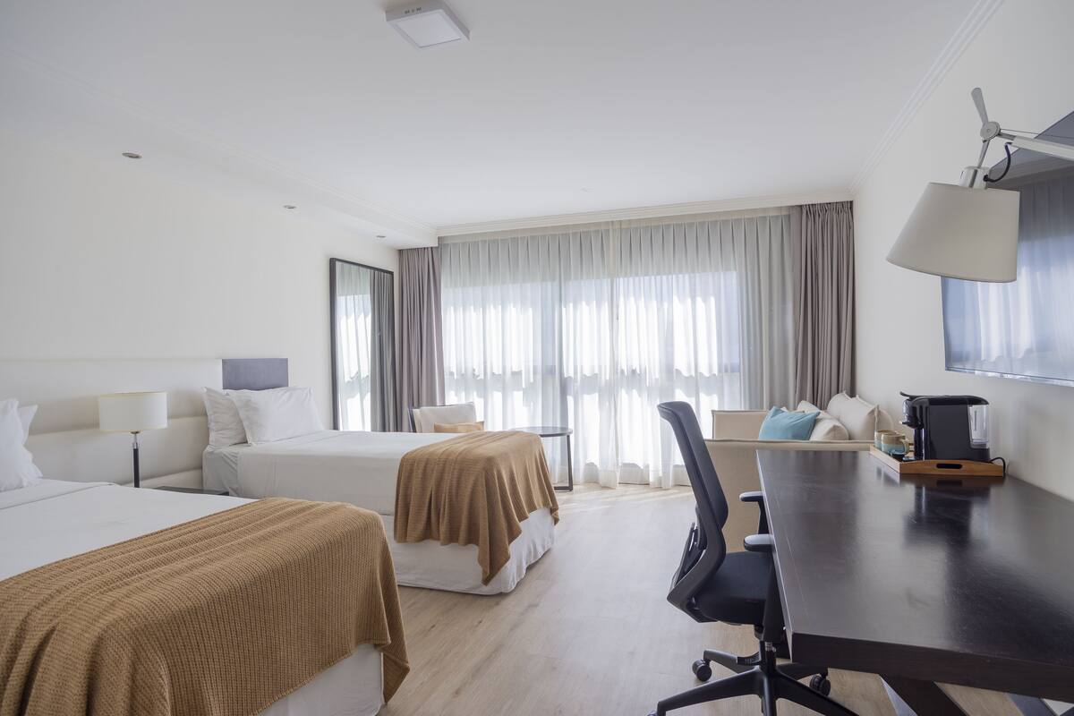 A hotel room with two beds, a desk, a chair, a coffee maker, and a large window with curtains.