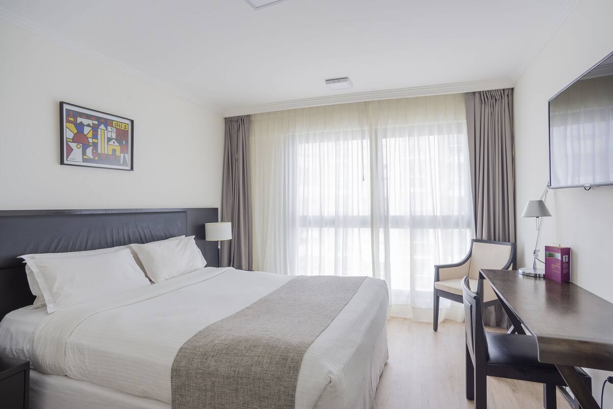 A hotel room with a large bed, a desk, a chair, a lamp, and a painting on the wall.