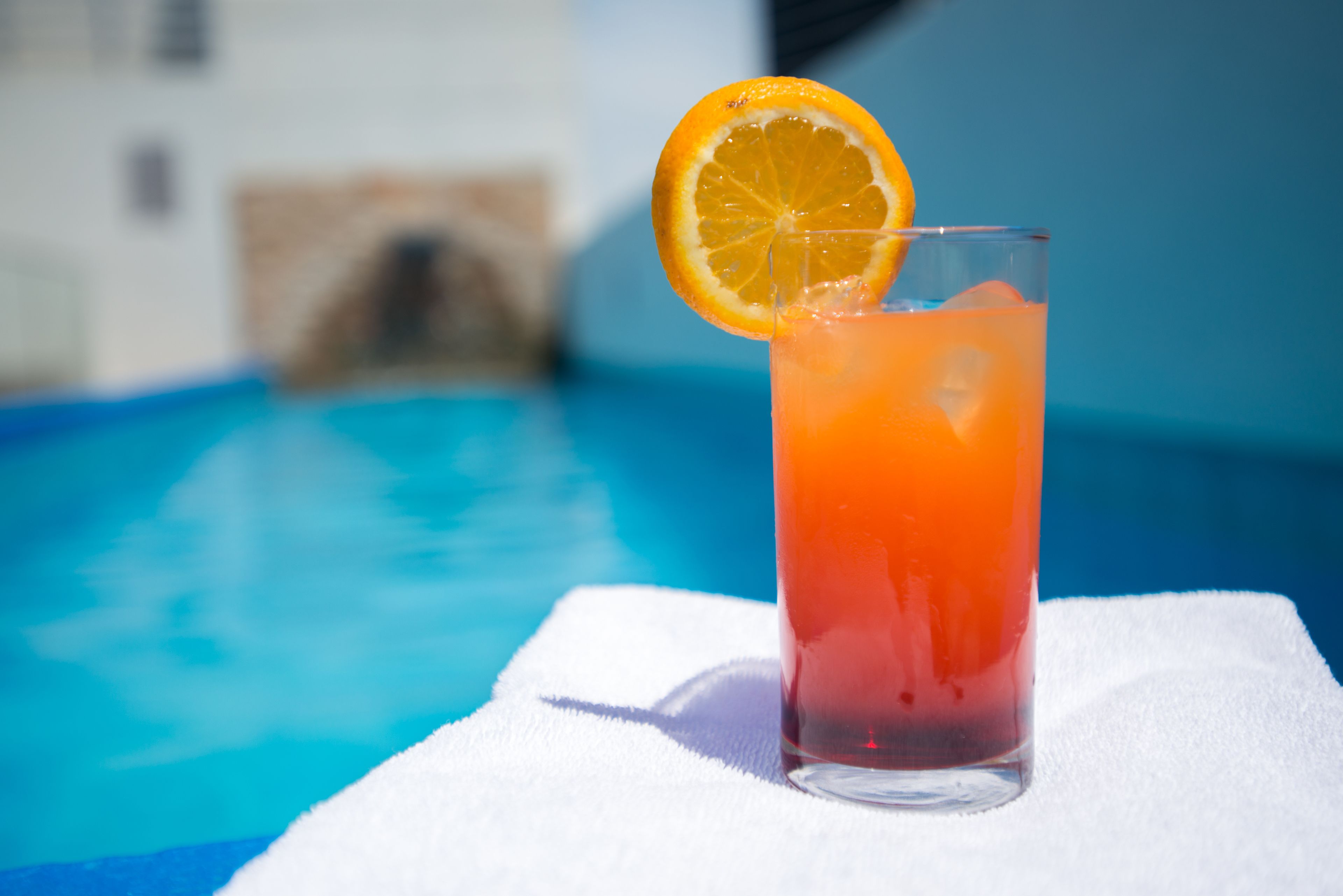 Un verre d’une boisson de couleur orangée, garni d’une tranche d’orange, posé sur une serviette blanche au bord d’une piscine.