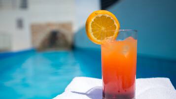 Un verre d’une boisson de couleur orangée, garni d’une tranche d’orange, posé sur une serviette blanche au bord d’une piscine.