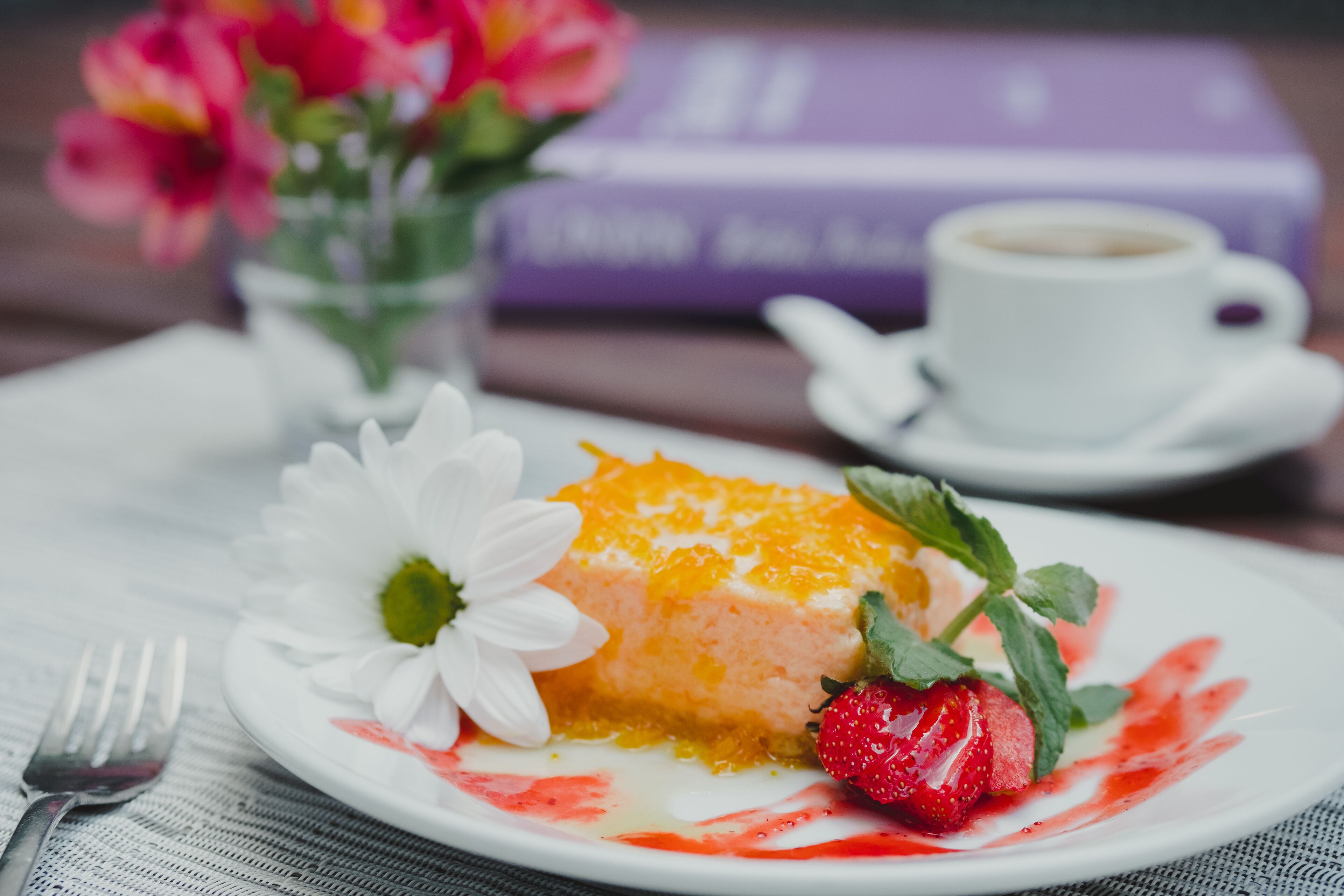 Un plato con postre, una rebanada de pastel, una fresa y una flor blanca sobre una mesa.
