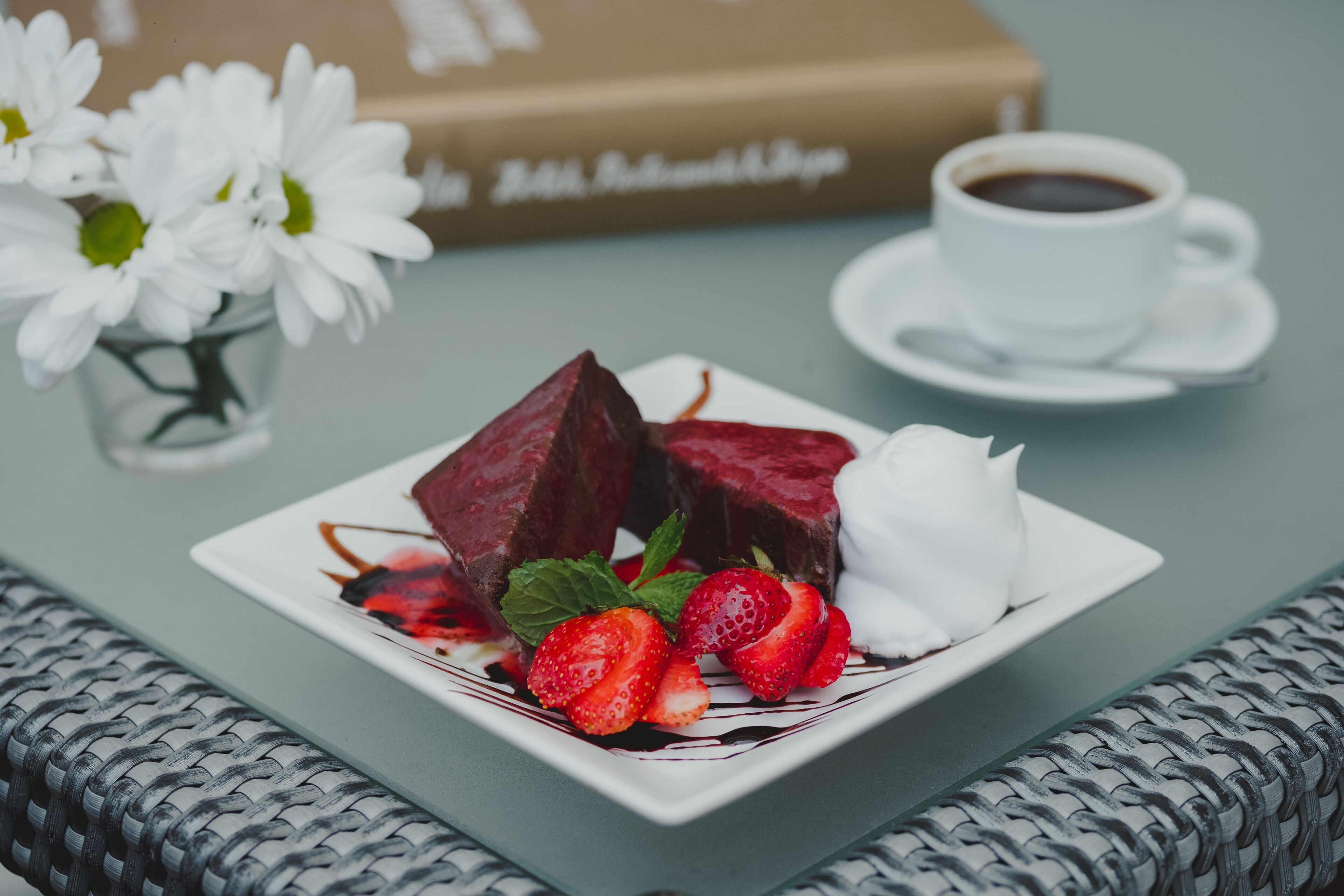 Ein Dessertteller mit Erdbeeren und einer weißen Nachspeise, eine Tasse Kaffee und ein Buch im Hintergrund.