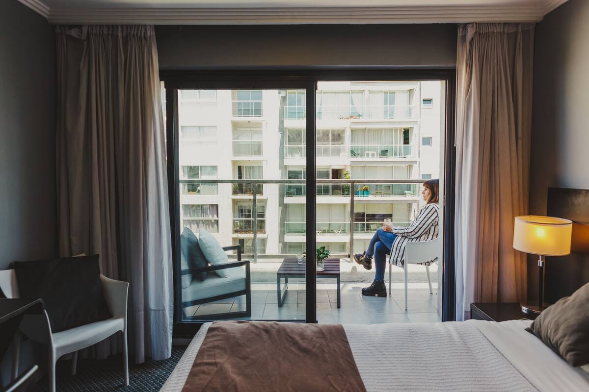 A hotel room with a bed, a desk, a chair, a table, and a view of a building through a glass door.