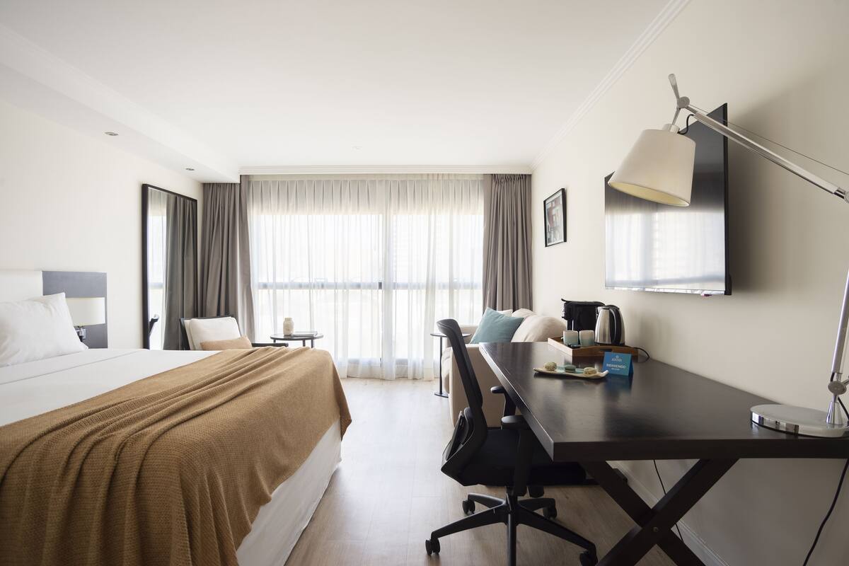 A hotel room with a bed, a desk, a chair, a TV, and a window with curtains.