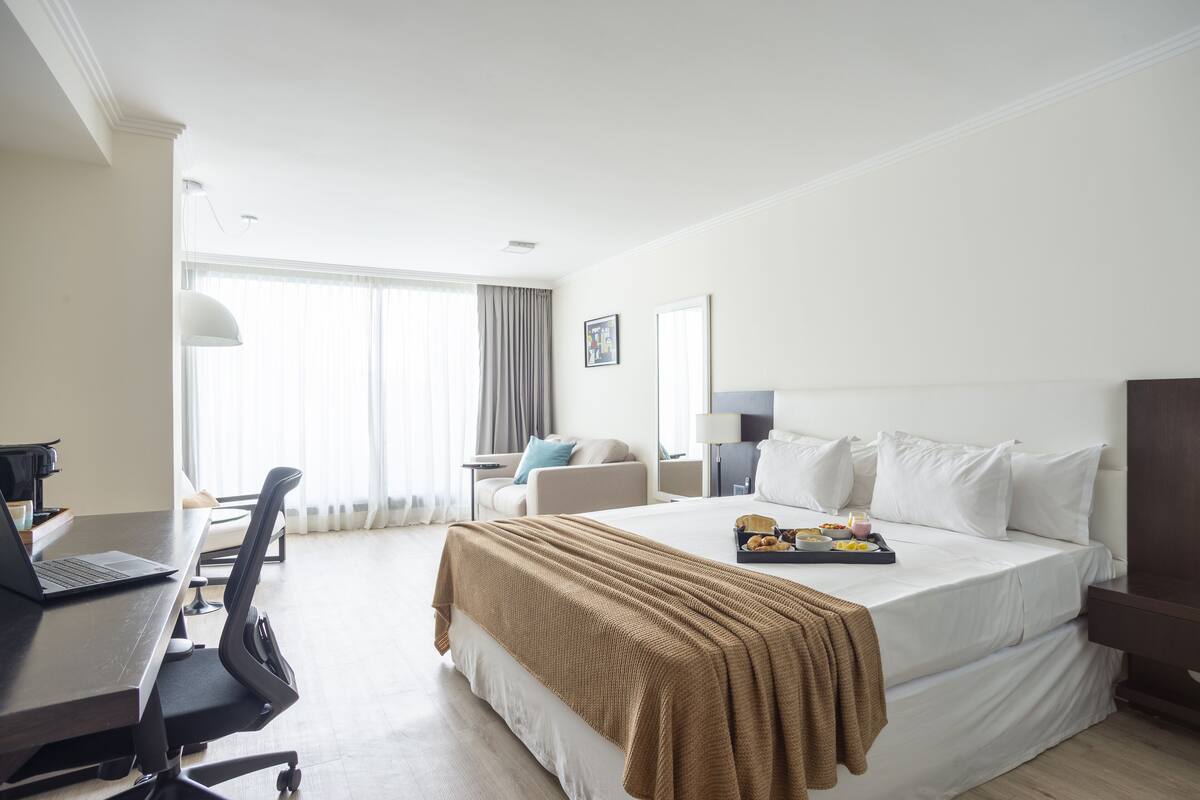A hotel room with a bed, a desk with a laptop, a chair, a bedside table with a lamp, and a window with curtains.