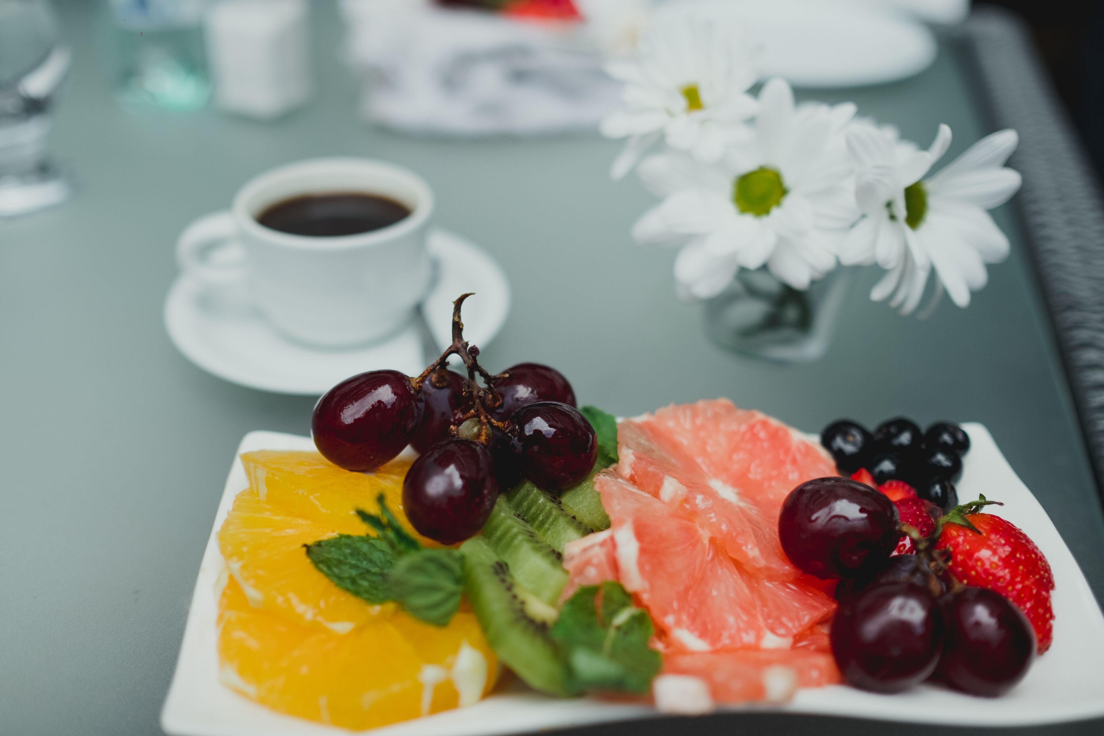 A breakfast setting with a plate of fruit, a cup of coffee, and a vase of white flowers on a table.