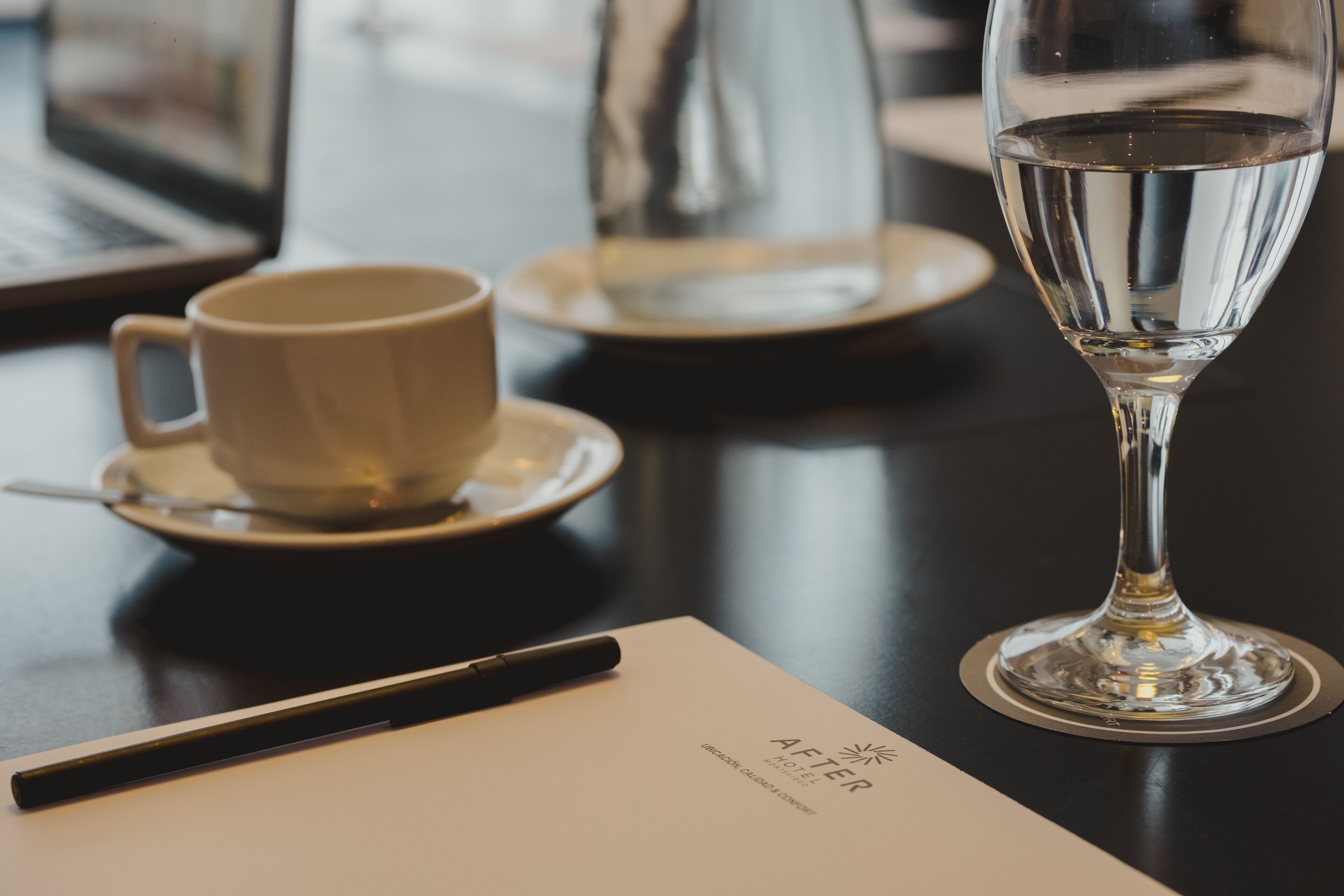 Una mesa con una taza blanca, un plato bajo y un vaso de agua. Un bolígrafo descansa sobre un papel.