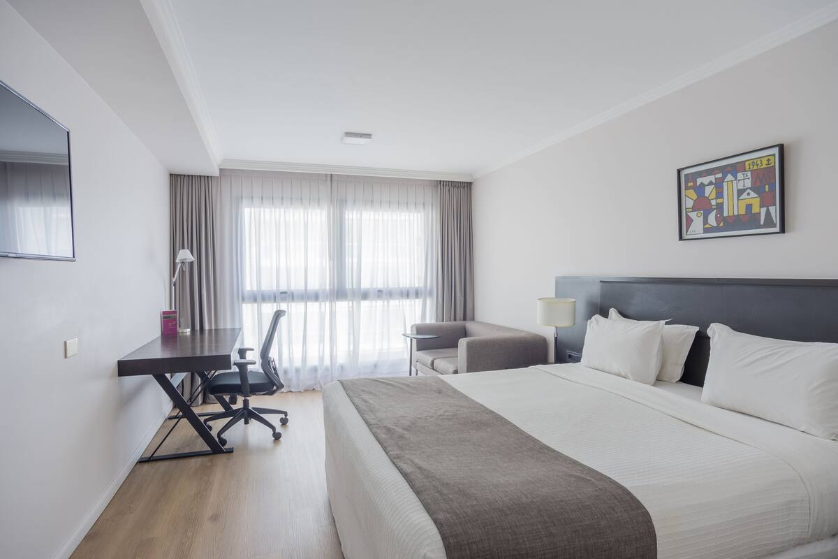 A modern hotel room with a large bed, a desk, a chair, and a window with curtains.