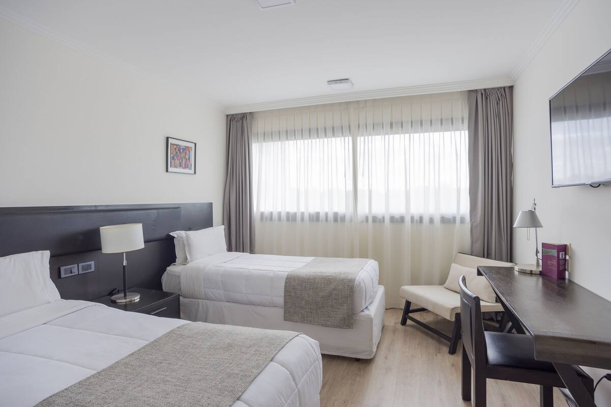 A hotel room with two beds, a desk, a chair, a lamp, and a large window with curtains.