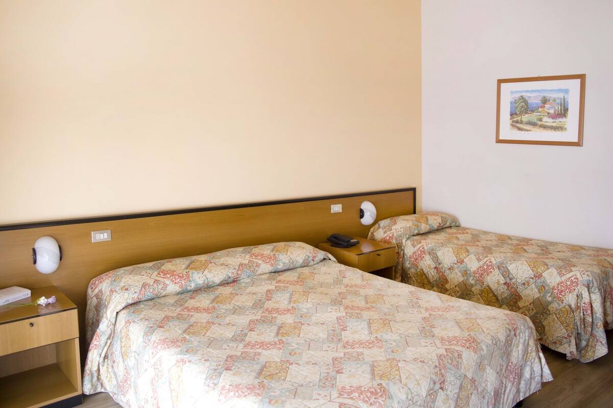 A hotel room with two single beds, wooden headboards, a bedside table with a lamp, and a framed picture on the wall.