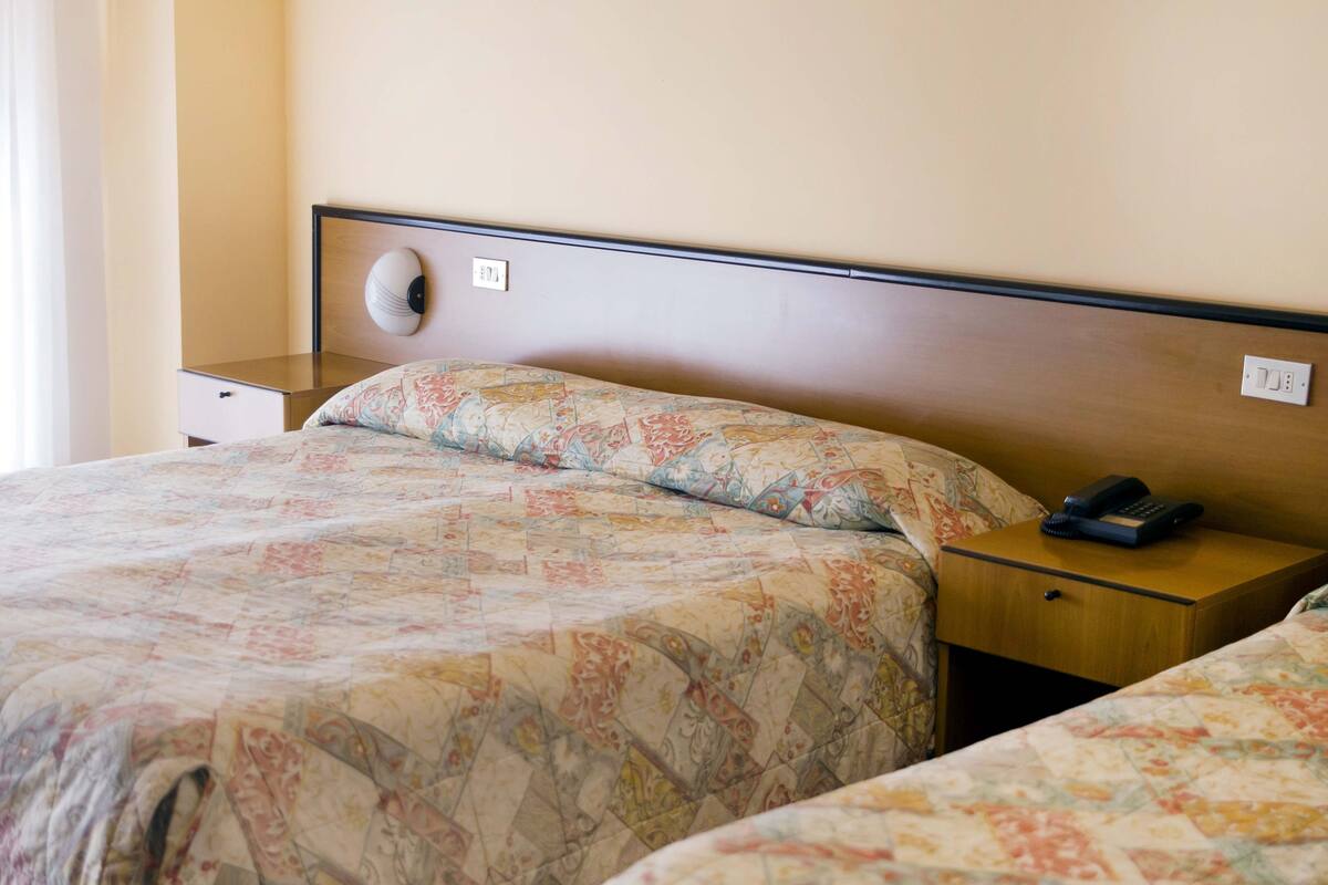 A hotel room with two beds, each with a floral-patterned bedspread, a wooden headboard, a bedside table with a phone, and a wall socket.