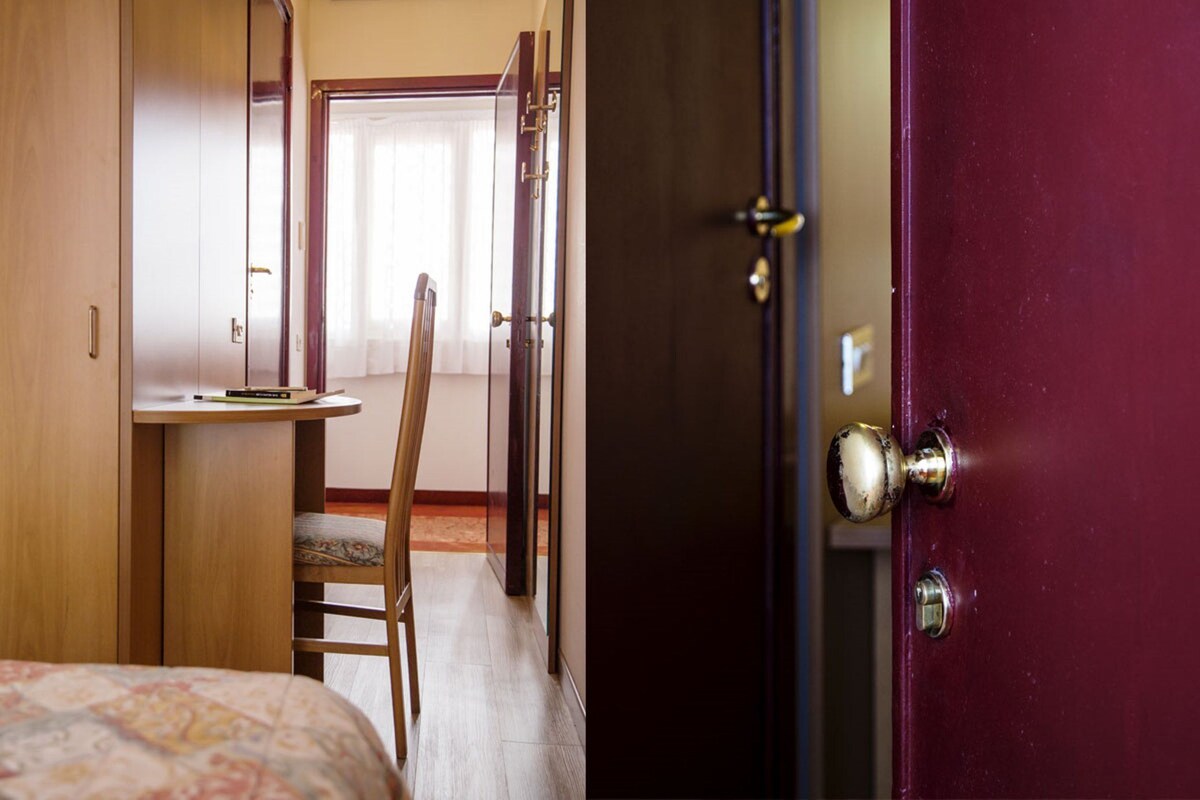 A hotel room with a bed, a desk, and a chair. There is a door with a handle and a lock.