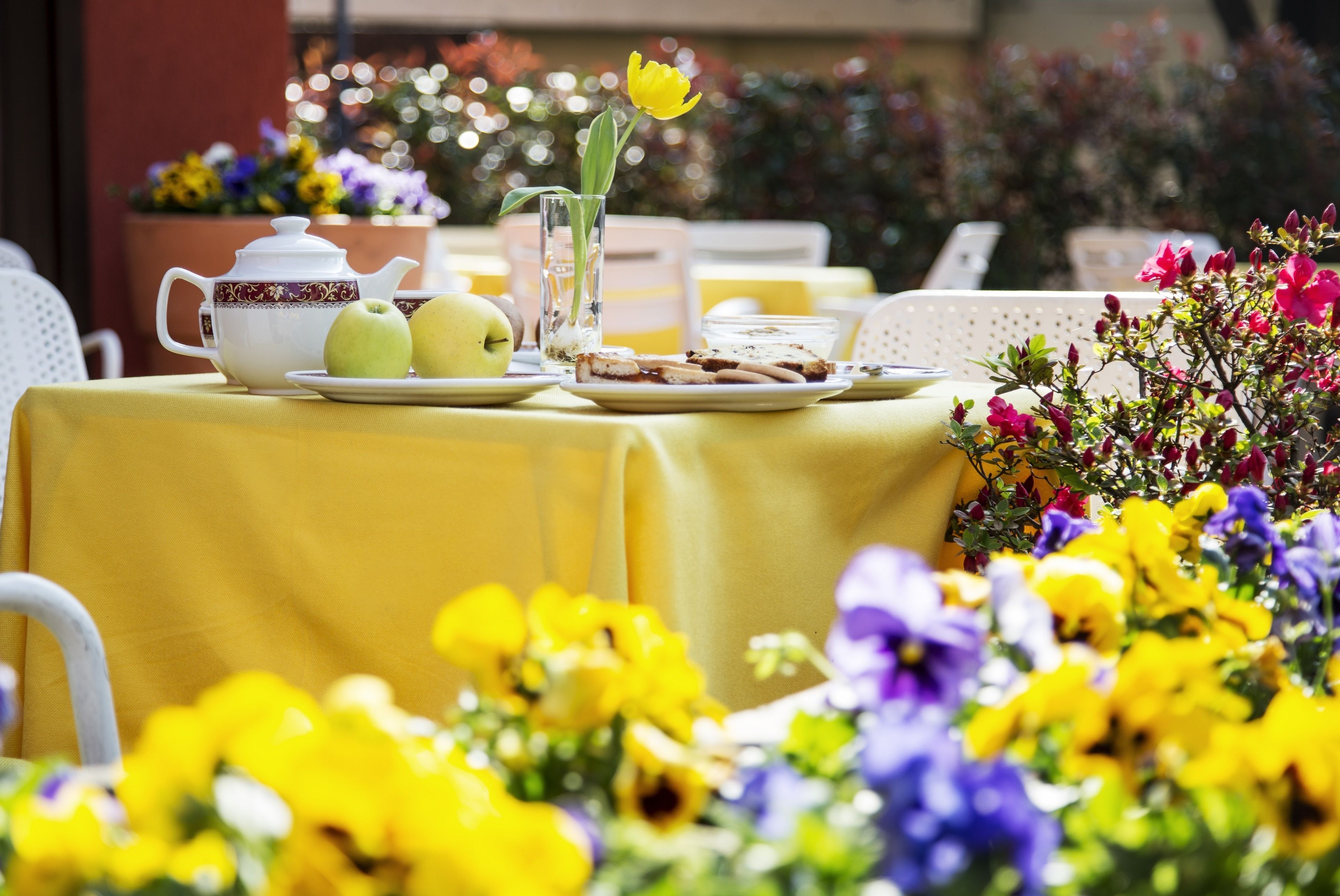 Een gedekte tafel met een theepot, appels, een glas water en een bord met eten, omringd door kleurrijke bloemen.