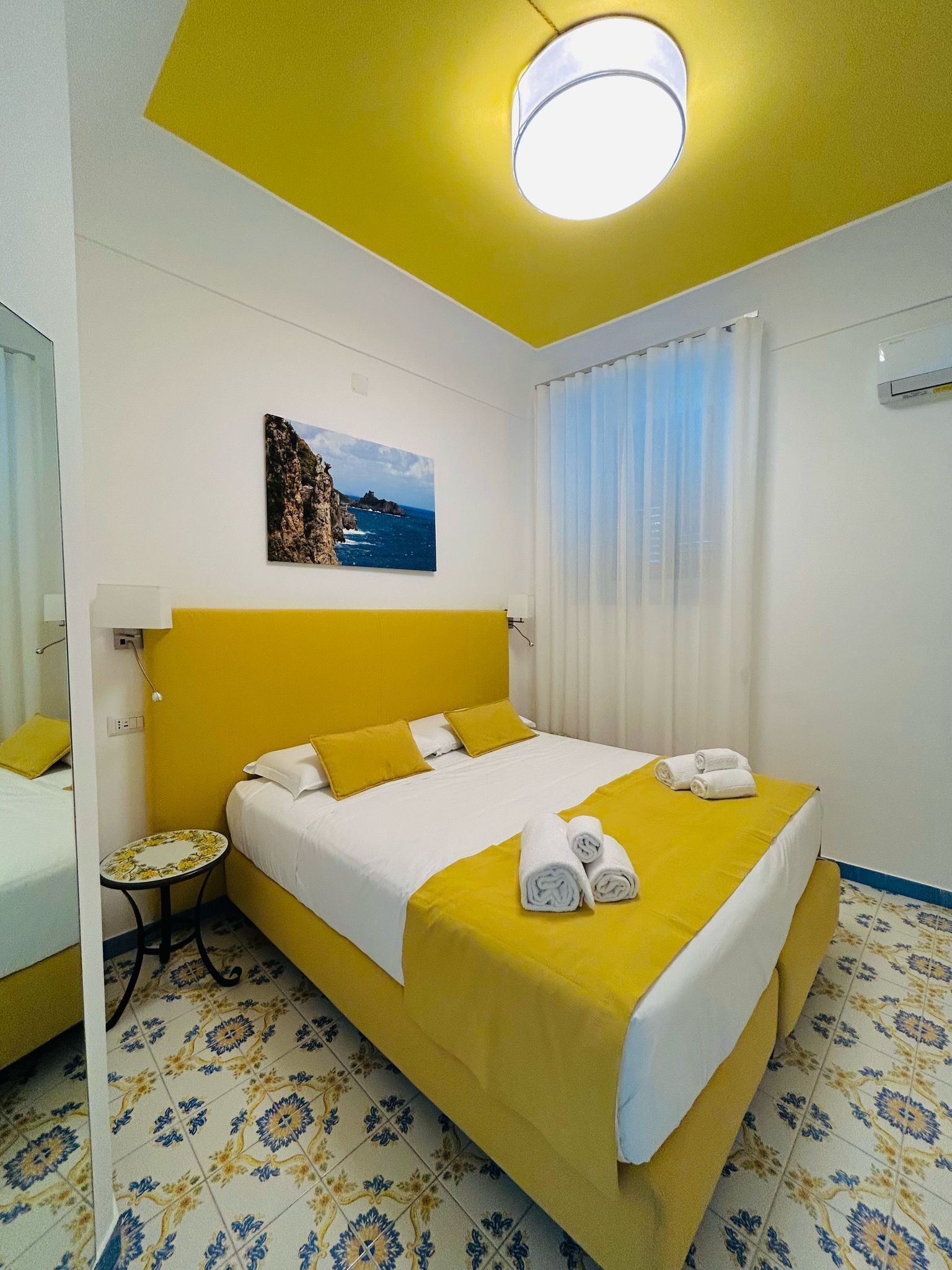 A hotel room with a bed, a small table, a painting, and a window with curtains.