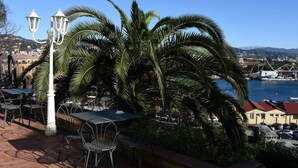 Ein Sitzbereich im Freien mit Tischen und Stühlen, einer Straßenlaterne und Blick auf einen Hafen mit Gebäuden und Hügeln im Hintergrund.