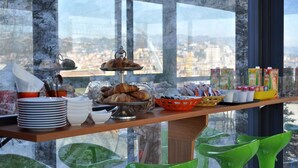 Ein Frühstücksbuffet mit Croissants, Gebäck und Orangensaftkartons auf einem Holztisch, mit Blick auf die Stadt durch große Fenster.
