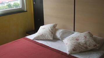 A hotel room with a bed, two pillows, a red blanket, a nightstand, and a lamp.