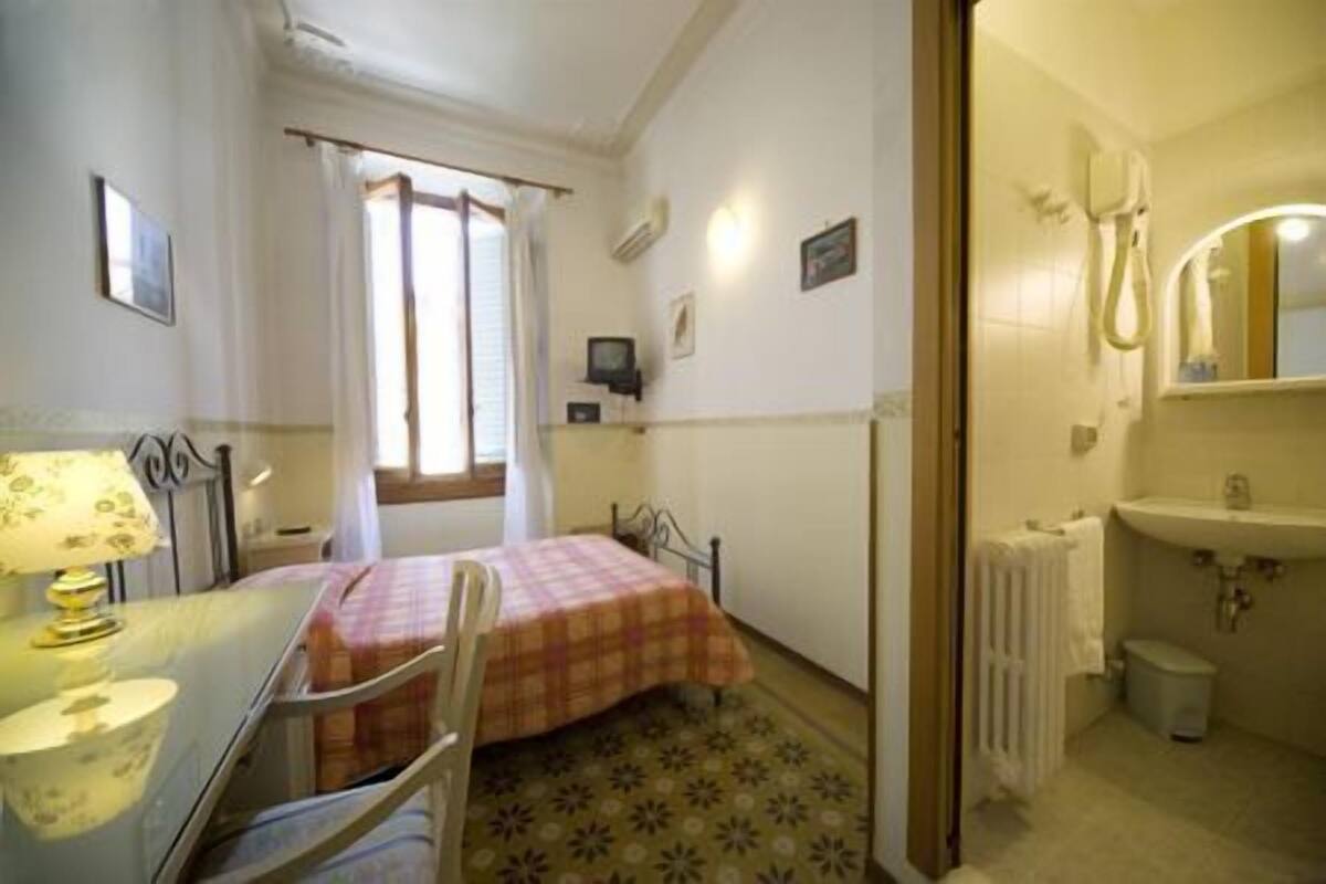 A small, cozy bedroom with a single bed, a desk with a lamp, a window with curtains, and a bathroom with a sink and mirror.