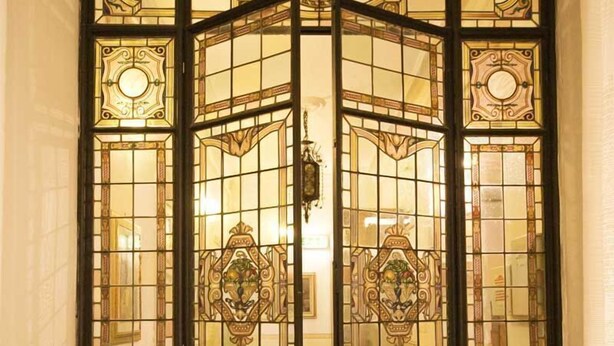 Una habitación con piso a cuadros, una puerta grande y ornamentada con paneles de vidrio y una pequeña mesa con un florero.