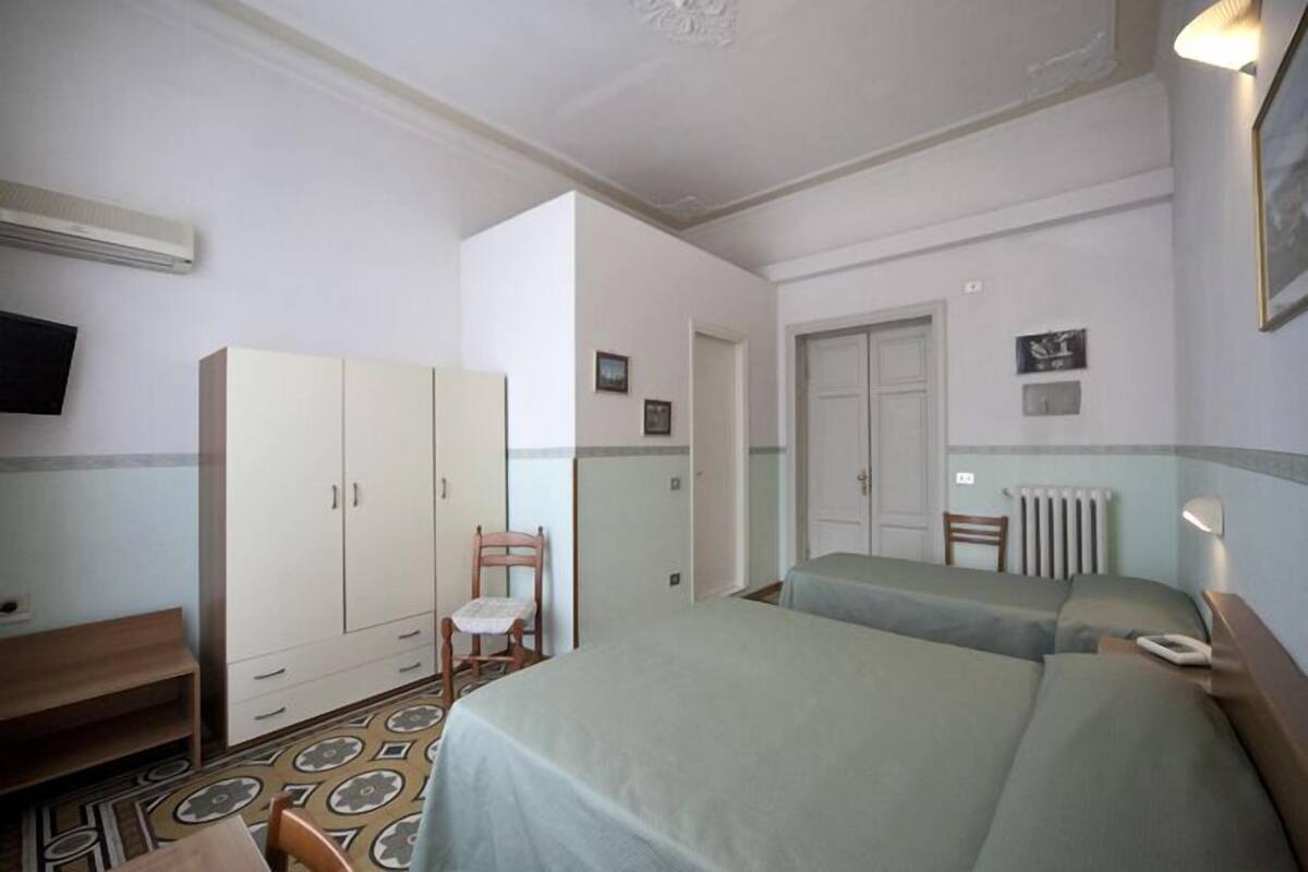 A hotel room with two beds, a wardrobe, a chair, a desk, and a radiator.